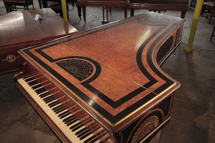 Piano lid is in burr walnut with black borders and carved decoration in low relief. At the piano tail, a circular motif bordered with spiralling rope motif contains four symmetrical anthemions. Above the piano fall, a semi-circular sun motif is bordered with stylised swags and palm leaves.
